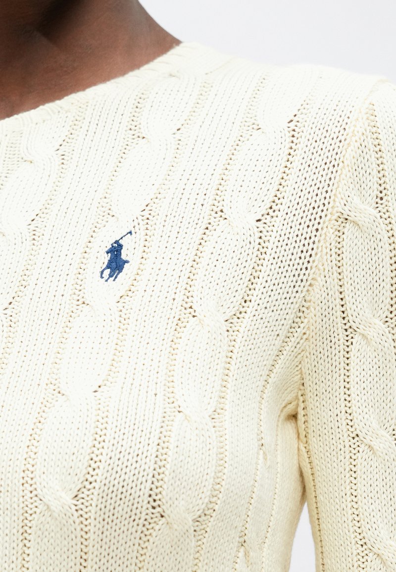 Pull en maille crème avec un col rond et un logo brodé bleu marine. La texture est douce avec des motifs de tricot visibles.