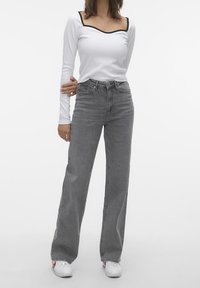 Camisa blanca de manga larga con ribete negro, combinada con pantalones vaqueros grises de pierna ancha y cintura alta. El modelo lleva zapatillas blancas.