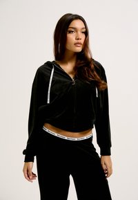 Cropped hoodie em veludo preto com fecho de zíper à frente, cordões brancos e uma cintura correspondente com o logo 'Juicy Couture'. Textura suave.