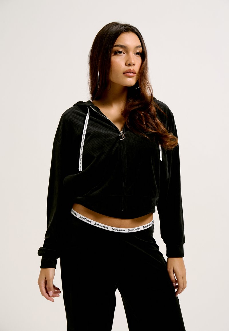 Cropped hoodie em veludo preto com fecho de zíper à frente, cordões brancos e uma cintura correspondente com o logo 'Juicy Couture'. Textura suave.