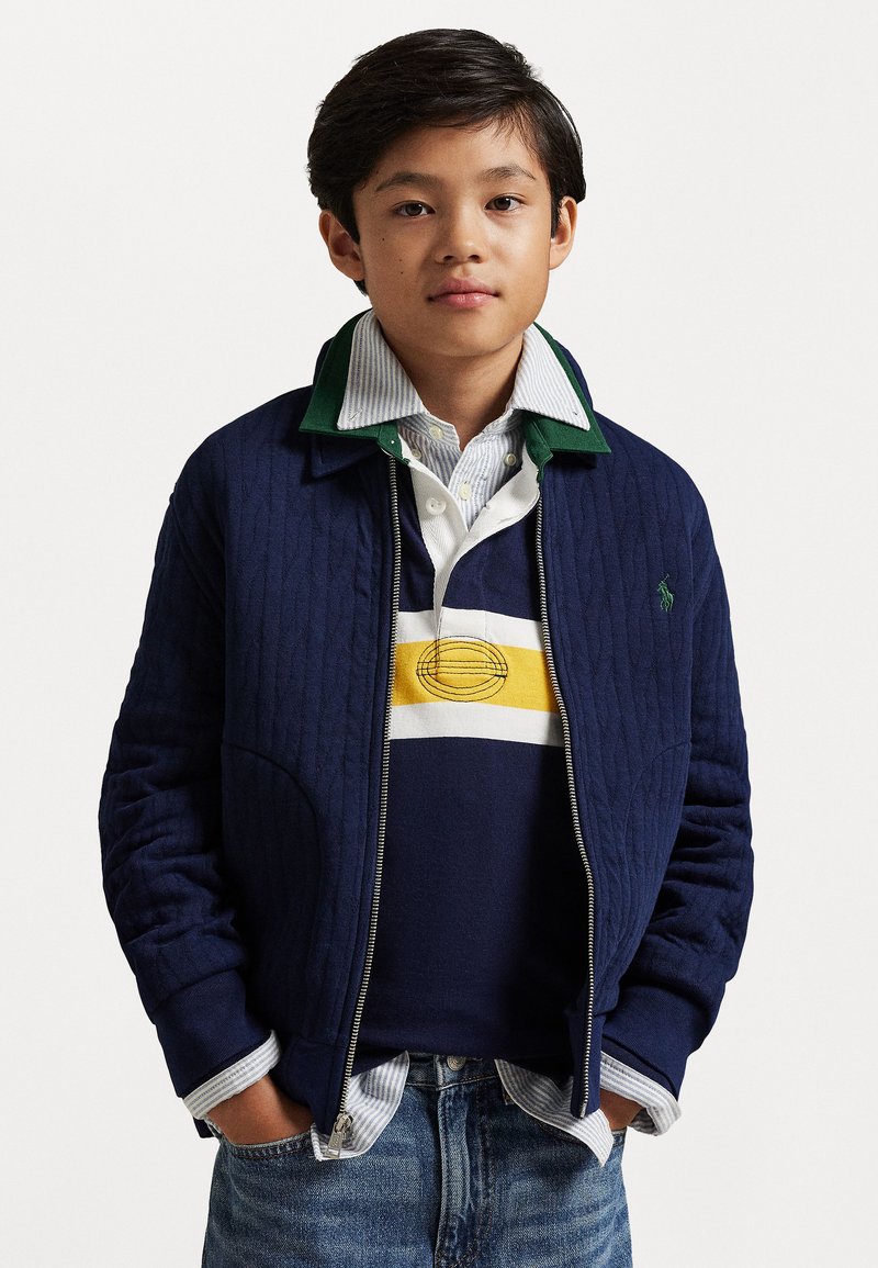 Jūras spēku zila quiltota jaka ar zaļu apkakli, uzvilkta virs svītraina polo krekla ar dzelteniem un jūras spēku zila akcentiem. Kombinēta ar gaiši ziliem džinsiem.