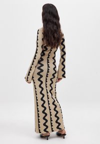 Langsleeve aansluitende jurk in beige breisel met zwart zigzagpatroon, met uitlopende mouwen en een vloerlange uitstraling.