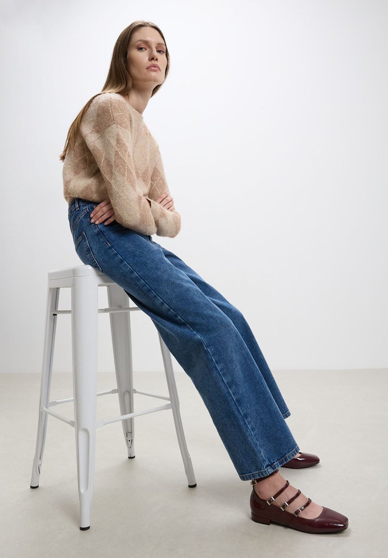 Maglione in maglia beige con motivo a rombi, jeans blu a gamba larga e scarpe in pelle bordeaux con accentuazioni a strappo, seduto su uno sgabello bianco.