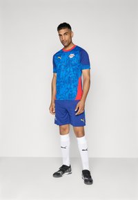 Maglia sportiva blu con motivo grafico; accenti laterali rossi. Abbinata a pantaloni blu, calze bianche e scarpini neri. Branding Puma visibile.