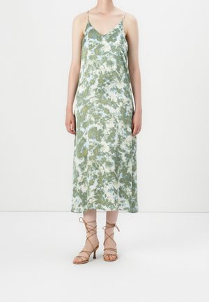 Vrouw die een enkellange, mouwloze tie-dye jurk met dunne bandjes en beige vetersandalen met hoge hakken draagt, staand op een witte vloer.