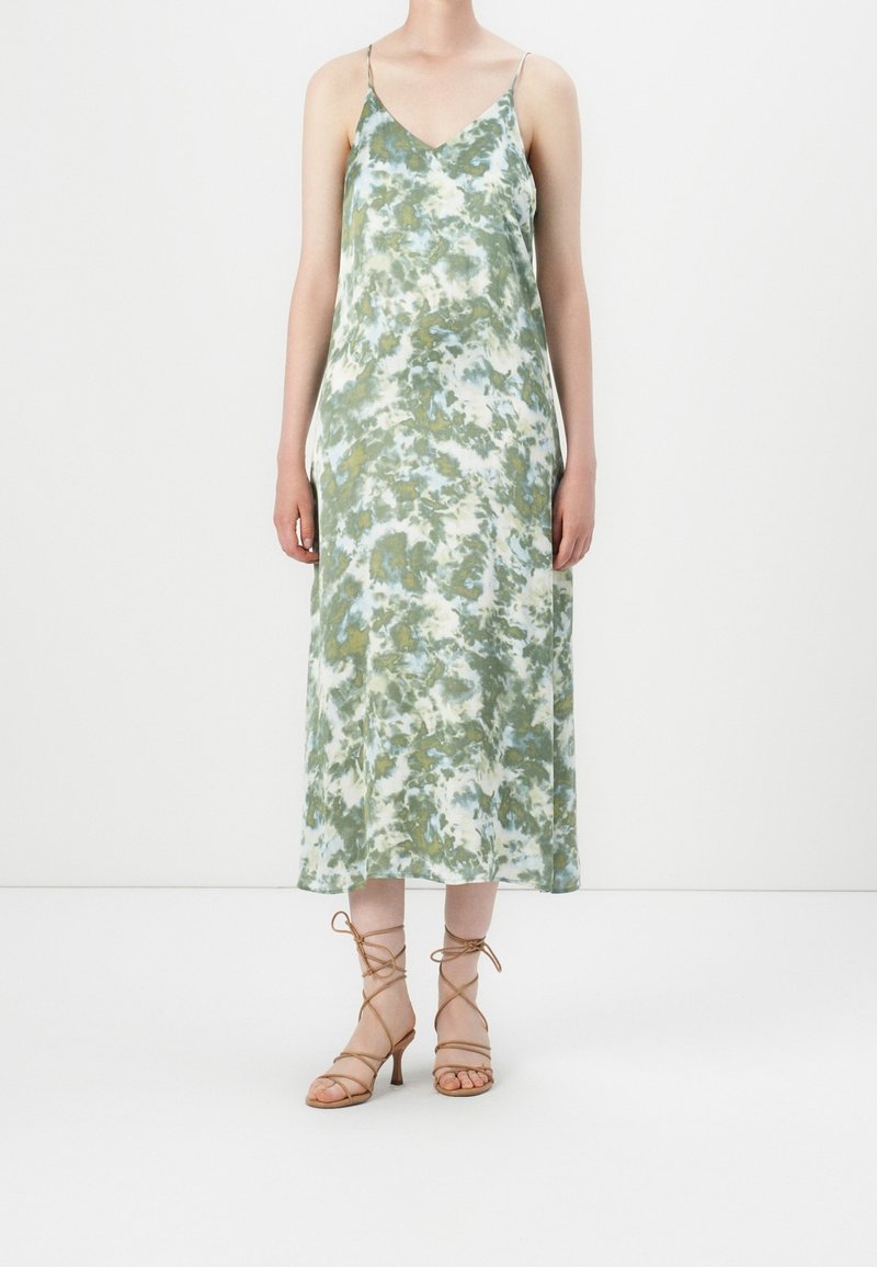 Femme portant une robe sans manches tie-dye longue jusqu'aux chevilles avec des fines bretelles et des sandales à talons hauts beiges à lacets, debout sur un sol blanc.