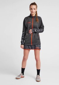 Graue Sportjacke mit orangefarbenem Reißverschluss, mit chevronmusterärmeln. Kombiniert mit einem passenden grauen Rock und sportlichen Schuhen.