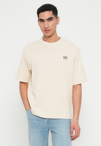Junger Mann, der ein beiges, übergroßes T-Shirt mit dem Text "THE HAZE JAZZ LOUNGE" auf der Brust trägt, kombiniert mit hellblauen Jeans, vor einem weißen Hintergrund stehend.