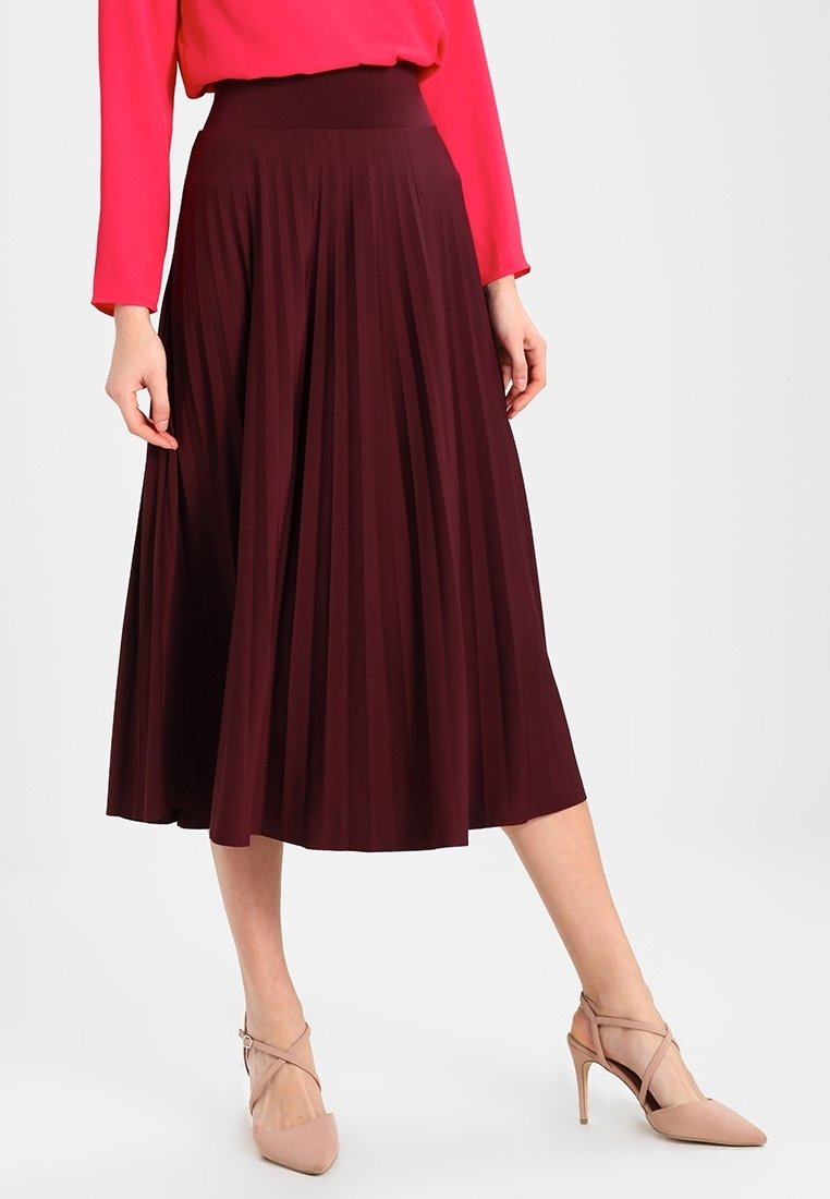 Femme portant une jupe midi plissée bordeaux avec une large ceinture, associée à des escarpins nude à bout pointu et un chemisier rose vif.