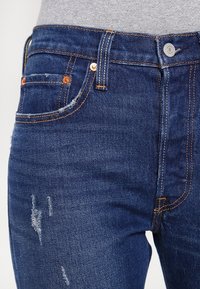 Los jeans de mezclilla azul oscuro presentan un ajuste Slim con remaches de cobre, secciones desgastadas y un diseño clásico de cinco bolsillos. La costura es de color marrón claro.