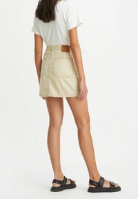 Žena stojící zády v béžové pruhované mini sukni, bílé tričko a černé sandály, na bílém pozadí.