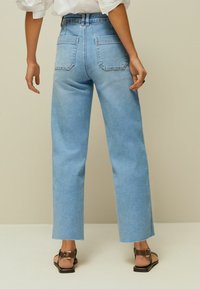 Jeans de mezclilla azul claro con cintura alta, corte recto y dos bolsillos traseros, combinados con sandalias planas marrones. Textura suave.