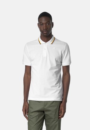 Giovane uomo in piedi che indossa una polo bianca con collo a righe gialle e nere e pantaloni verde oliva su sfondo uniforme.