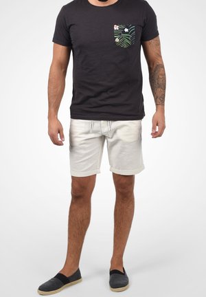 Ciemny T-shirt z kieszonką zdobioną wzorem tropikalnych liści, połączony z jasnymi szortami i szarymi butami wsuwanymi.