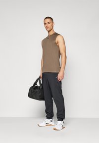 Top sin mangas marrón, pantalones deportivos negros y zapatillas blancas con detalles en negro. La modelo sostiene un bolso de viaje negro. Diseño simple y funcional.