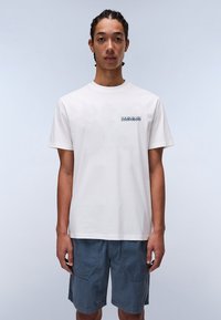 Hvid bomulds t-shirt med rund hals og logo på brystet. Modellen har blå shorts på og står foran en lysegrå baggrund.