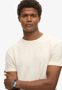 Lichtbeige t-shirt met korte mouwen en een ronde hals, met een klein logo op de mouw. Het heeft een gladde stof en een relaxte pasvorm.