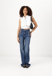 Top bianco cropped senza maniche con righe verticali; jeans in denim blu a vita alta con più bottoni dorati sui lati; borsa nera.