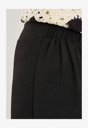 Pantalones negros texturizados con cintura elástica y bolsillo delantero, combinados con un top de crochet crema con detalle de pompones negros.