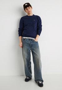 Νέος άνδρας στηριγμένος σε τοίχο φορώντας ναυτικό μπλε φούτερ, φαρδιά ξεβαμμένα τζιν, μαύρα αθλητικά παπούτσια και σκούρο καπέλο, με το ένα χέρι στην τσέπη.