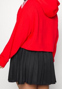 Röd kort hoodie med vita randiga detaljer på axlarna, kombinerad med en svart plisserad kjol. Tygerna verkar mjuka med en slät yta.