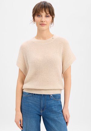 Frau mit kurzen braunen Haaren trägt einen beigen kurzärmligen Strickpullover und blaue hoch taillierte Jeans, steht vor einem weißen Hintergrund.
