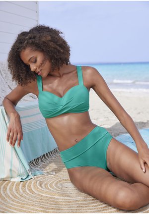 Türkisfarbenes Zweiteiler-Bikini mit einem gewundenen Design-Oberteil mit breiten Trägern und hochgeschnittenem Höschen. Glatte Textur, getragen in einer sandigen Strandkulisse.