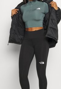 Schwarze Pufferjacke geöffnet, die ein kurzes teal-farbenes Top mit Stehkragen und hochgeschnittene schwarze Leggings mit weißem Logo zeigt. Glatte Stofftextur.