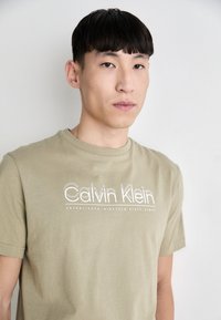 Olivově zelené bavlněné tričko s bílým logem "Calvin Klein", kulatým výstřihem, krátkými rukávy a jemnou texturou.