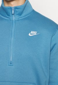 Sudadera Nike de color azul claro con un alto cuello, cierre de medio zip en la parte frontal, bordes acanalados y un logotipo bordado en blanco en el lado izquierdo del pecho. Textura de tela suave.