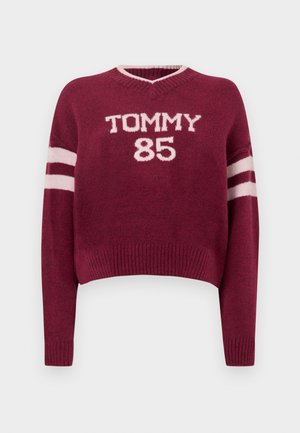 Bordo krāsas cropped džemperis ar V veida izgriezumu, uz tā baltā krāsā rakstīts "TOMMY 85" un uz piedurknēm ir balti svītraini akcenti. Mīksta adīta tekstūra.