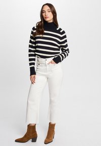 Schwarz-creme gestreifter Pulli mit hohem Kragen, kombiniert mit weißen hoch taillierten Jeans und braunen Wildleder-Stiefeletten mit Blockabsatz.