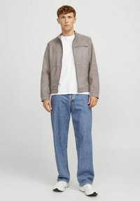 Giacca in suede grigia con colletto alto e tasca con zip, abbinata a una t-shirt bianca e jeans in denim blu, indossata con sneaker di colore chiaro.