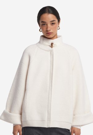 Kvinde iført en hvid tekstureret kappe med brede ærmer og en høj krave, der er fastgjort med en træknap.