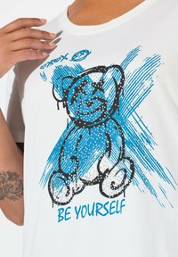 Bílá bavlněná tričko s modrou grafikou medvěda včetně černých akcentů, zdobením korálky a textem "BUĎ SÁM SEBOU" pod designem.