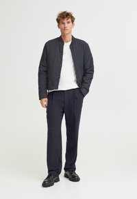 Giacca bomber nera trapuntata con colletto a coste, indossata sopra una t-shirt bianca, abbinata a pantaloni blu navy e scarpe nere, su uno sfondo semplice.