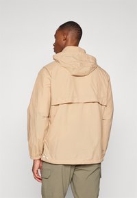 Veste à capuche beige clair avec une coupe ample, des poignets élastiques et un détail d'aération dans le dos. Fabriquée à partir d'un matériau léger et texturé.