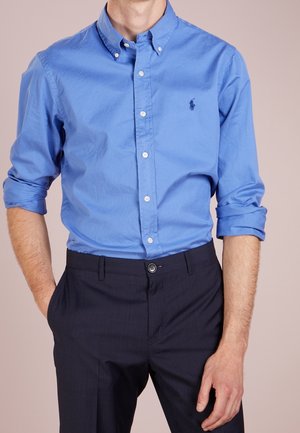 Mies, joka pukeutunut siniseen napitettuun paitaan, jossa hihat käärityt ylös, ja tummiin räätälöityihin housuihin, kädet rentoina lantion kohdalla.