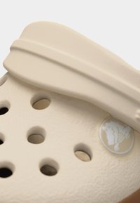 Μπεζ λαστιχένια κλογκς με οπές αερισμού και ρυθμιζόμενο λουράκι, με ένα διακριτικό ανάγλυφο λογότυπο κοντά στην περιοχή της φτέρνας.