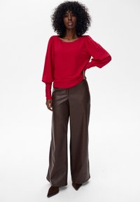 Kobieta z kręconymi włosami, ubrana w czerwoną bluzkę z długim rękawem i szerokie brązowe skórzane spodnie, stoi z jedną ręką na biodrze.