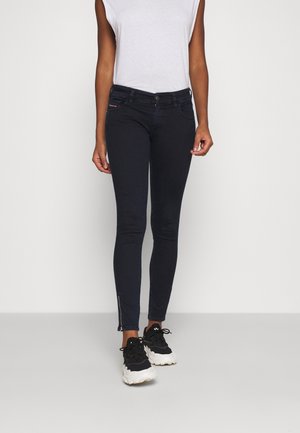 Personne portant un top blanc sans manches, un jean skinny foncé avec des fermetures éclair aux chevilles, et des baskets noires épaisses, debout devant un fond blanc.