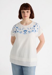 Blusa blanca con bordado floral azul en el escote. Mangas cortas enrolladas, diseño en capas y tela ligera.