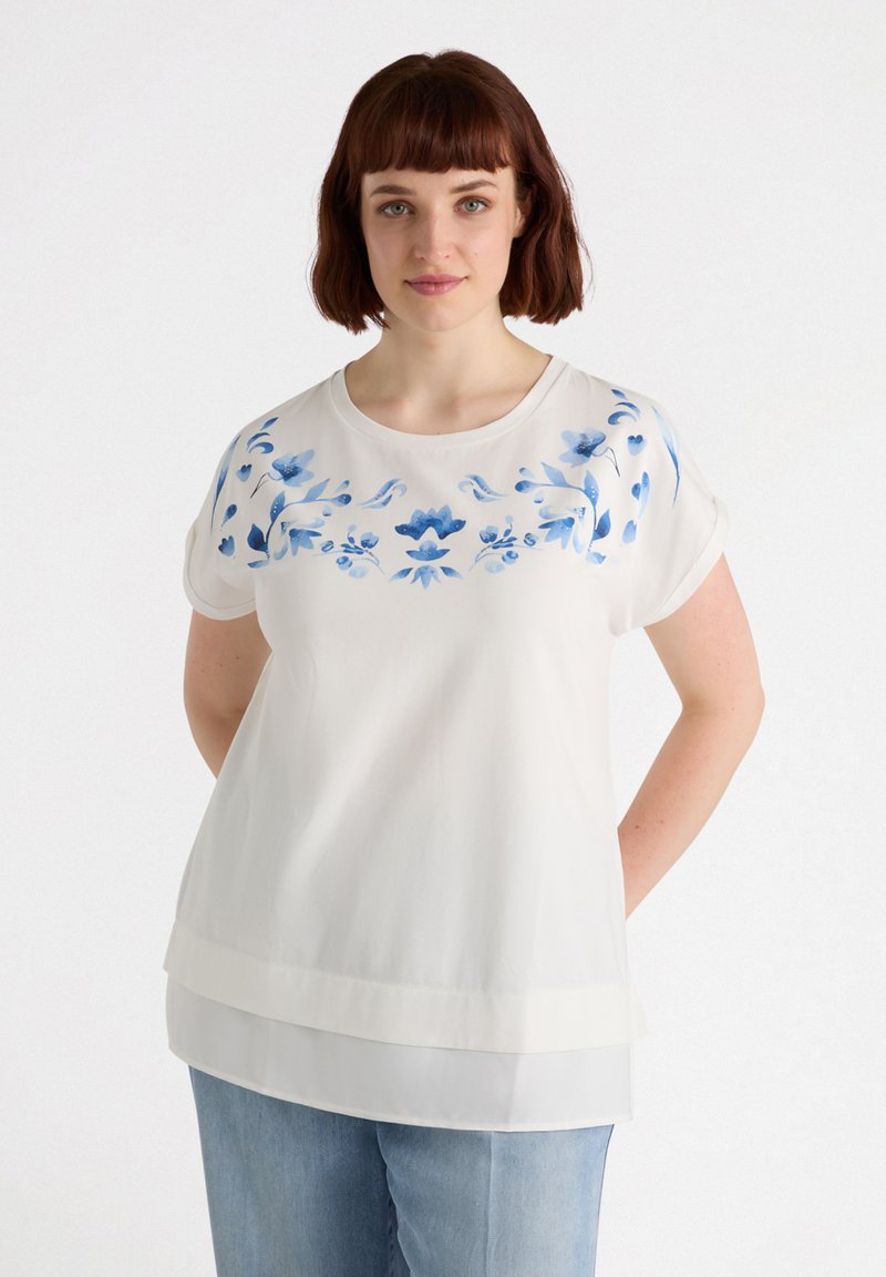 Blusa blanca con bordado floral azul en el escote. Mangas cortas enrolladas, diseño en capas y tela ligera.