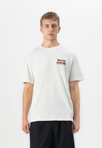 Ung mann iført en hvit Rip Curl Australia t-skjorte og svarte shorts, stående mot en enkel hvit bakgrunn.
