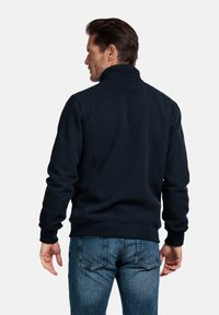 Marineblauwe gestructureerde sweatshirt met hoge kraag, ribgebreide manchetten en zoom. Draagt het met lichtblauwe jeans, die een casual achterkant laten zien.