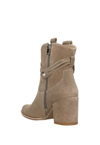 Béžové semišové kotníkové boty s kulatou špičkou, bočním zipem a dekorativními pásky; mají robustní blokový podpatek a hladký povrch.