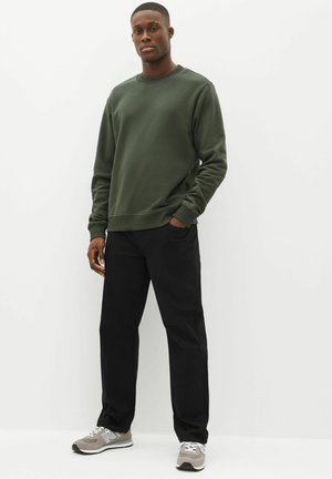 Mann steht vor weißem Hintergrund, trägt einen dunkelgrünen Pullover, schwarze Hose und graue Turnschuhe, eine Hand in der Tasche.