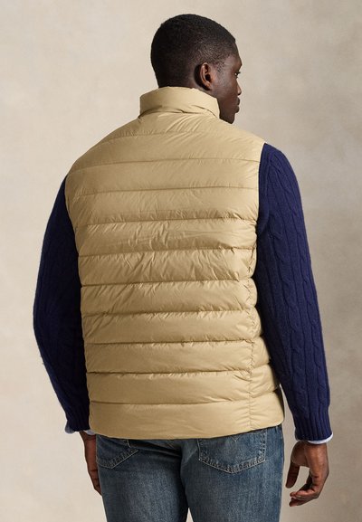 Bēša izolēta veste ar augstu apkakli, ar teksturētām tumši zila kabeļu adījuma džempera piedurknēm un zilām džinsu biksēm. Gluda apdare un šūts dizains.