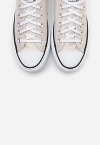 Béžové plátěné tenisky s bílou gumovou podrážkou a černými akcenty. Mají bílé šňůrky a klasické špičky. Hladký povrch a zaoblený tvar.