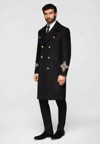 Schwarzer Wollmantel mit goldenen Knöpfen und bestickten Blumenakzenten an den Ärmeln. Maßgeschneiderte Passform, doppelreihiges Design, kombiniert mit schwarzer Hose.